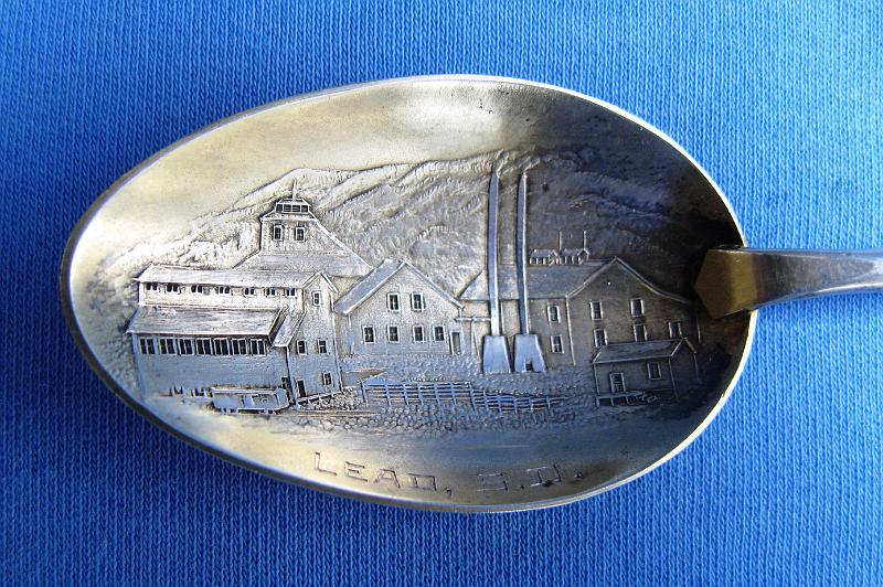 Souvenir Mining Spoon Bowl Lead SD.JPG - SOUVENIR MINING SPOON LEAD SD HOMESTAKE MINE - Sterling silver spoon, 6 1/4 in. long, embossed mining scene of Homestake Mine in bowl with engraved LEAD, S. D. in bowl, handle with mining pan, picks and rope  [The founding of Lead, SD and the discovery of the Homestake Gold Mine are intertwined as one of the earliest enterprises associated with the Gold Rush of 1876 in the northern Black Hills of what was then Dakota Territory.  Brothers Fred and Moses Manuel and their partner Hank Harney located their Homestake claim on April 9, 1876.  Moses Manuel liked what he saw in an outcropping of a vein of ore, referred to as a lead and pronounced "leed." The name stuck.  Soon more prospectors materialized, and no time was lost in selecting a site for a new town. On July 10, 1876 work began on laying out the town lots, and that work was completed the next day. Miners were offered the lots, 50 x 100 feet, but were required to build on the lots in 60 days or forfeit them. That spurred many to build on the front half and then sell the back half. Progress came quickly. Telegraph service began December 1st and by early 1877 four hotels, a grocery store, saloon, bakery and butcher shop were up and running.  In June 1877 George Hearst, who had earlier sent an agent to offer a bond to owners of the Homestake claim, bought the four and one half acre claim for $70,000. No stranger to mining, Hearst had mining interests in Missouri, California during its gold rush, Nevada, Utah, and Montana. He later represented the State of California in the United States Senate. He and his wife Phoebe had one son, William Randolph Hearst, who, rather than continue in his father's footsteps in the mining businesses, chose to manage his father's newspaper, the San Francisco Examiner. William became a publishing magnate and was a pioneer in the radio and television industries.  With a population of 8,392 in 1910, Lead was the second largest community in South Dakota. The employment opportunities for not only miners, but also laborers and mechanics were excellent. After George Hearst's death in 1891, his widow Phoebe made substantial contributions to the educational and cultural life of Lead.  She was responsible for the establishment of the first kindergarten in the entire West. In addition, she arranged for the Homestake Mining Company Homestake Opera House and Recreation Building to be constructed as gifts to the community from the company.  In 1974, most of Lead was added to the National Register of Historic Places under the name of the "Lead Historic District". Over four hundred buildings and 580 acres were included in the historic district, which has boundaries roughly equivalent to the city limits.  After more than 125 years of continuous operation, the Homestake Mine was the largest, deepest (8,240 feet) and most productive gold mine in the Western Hemisphere before closing in January 2002. Lead and the Homestake Mine have been selected as the site of the Deep Underground Science and Engineering Laboratory, a proposed NSF facility for low-background experiments on neutrinos, dark matter, and other nuclear physics topics, as well as biology and mine engineering studies.]